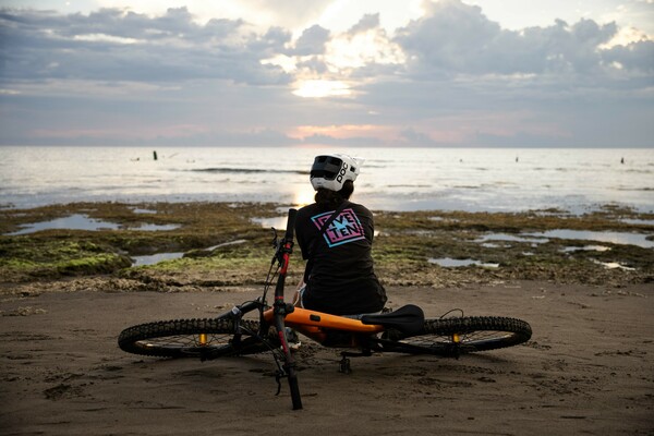 Samantha is sitting on the ground, her bike is lying next zu her and she looks out to sea in the distance